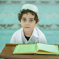 Young boy in traditional attire reading the Quran with a focused expression.