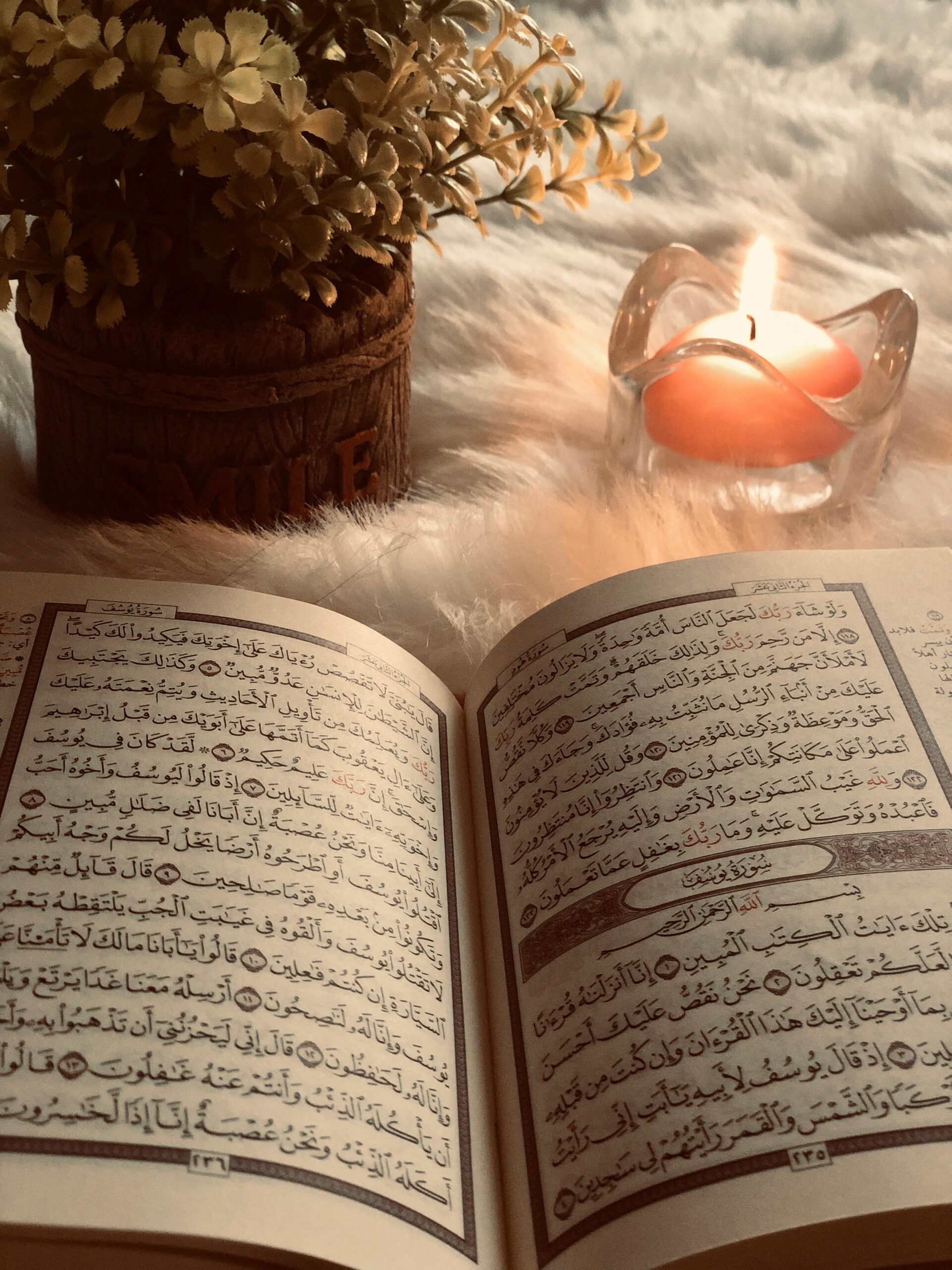 A beautifully open Quran placed beside a candle and flowers on a soft surface.